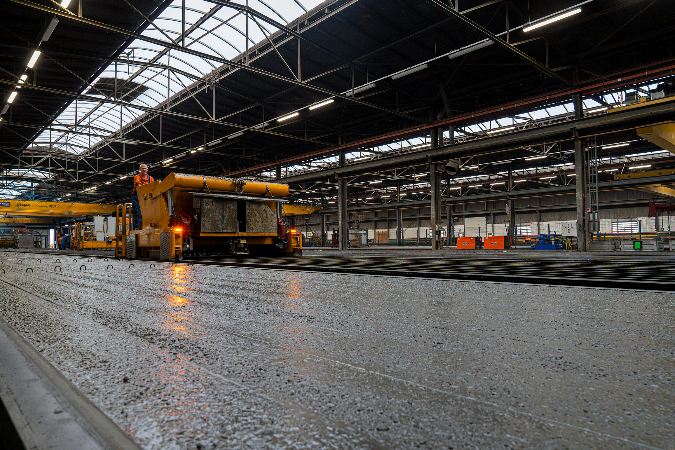 Een medewerker staat op een grote gele machine in een ruime fabriekshal met metalen dakconstructie en natuurlijk licht.