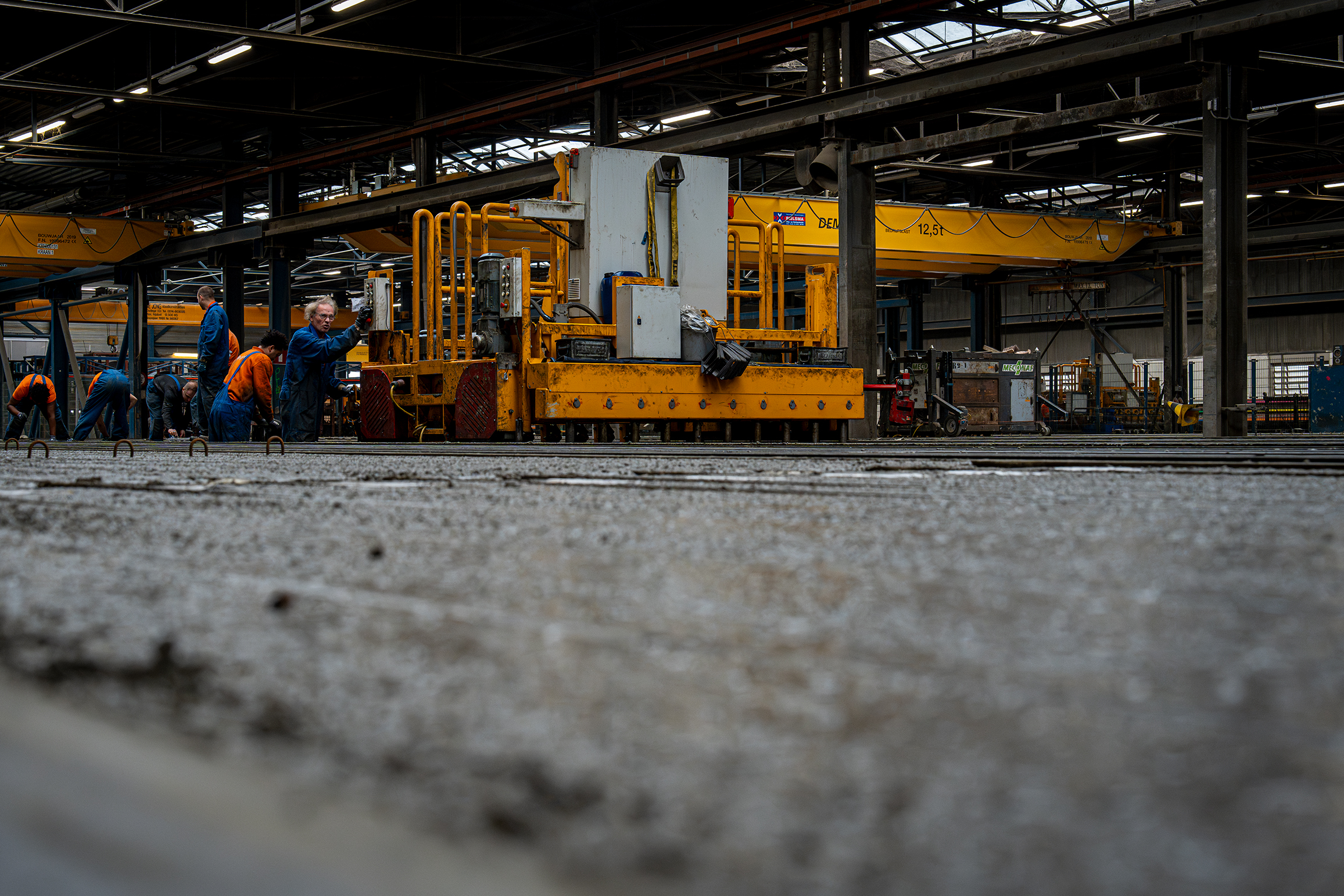 Werknemers in blauwe en oranje werkkleding bedienen een grote gele machine in een industriële fabriekshal met betonnen vloer en bovenloopkranen.