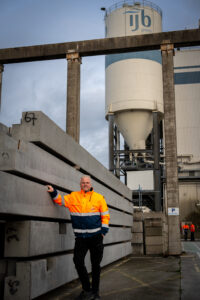 Een medewerker in oranje-blauwe veiligheidskleding leunt tegen een stapel betonnen liggers met op de achtergrond een silo met het logo van IJB Groep.